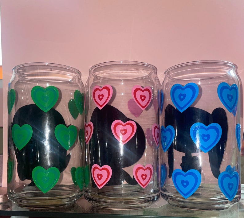 May include: Three clear glass cups with black silhouettes and colorful heart designs. The cups have green, pink, and blue hearts.