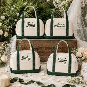 Puede incluir: Cuatro bolsos de lona color blanco roto con bordes y asas de color verde oscuro. Cada bolso presenta un nombre personalizado bordado en cursiva verde oscuro: Julia, Anna, Lucky y Emily. Los bolsos tienen una forma redondeada y estructurada.