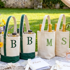 May include: Four off-white canvas wine bags with dark green trim and handles. Each bag features a large embroidered letter on the front: two bags with the letter B, one with M, and one with S. The bags are standing on a white lace table runner.
