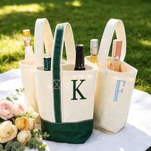 May include: Three off-white canvas wine tote bags with sturdy handles. One bag features a dark green base and handle trim with a dark green embroidered letter K. Another bag displays a light blue embroidered letter F. Each bag holds a glass bottle.