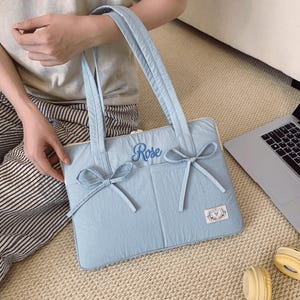 Peut inclure: Une sacoche d'ordinateur matelassée bleu clair avec deux longues bandoulières, deux nœuds en tissu décoratifs sur le devant et le mot Rose brodé en écriture bleue. Une petite étiquette rectangulaire avec un motif est fixée dans le coin.