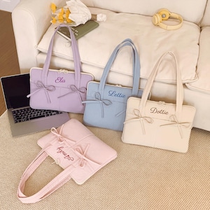 May include: Four rectangular laptop bags in pastel lavender, light blue, cream, and soft pink. Each bag features two top handles, a zippered closure, and two decorative bows on the front. The names Ela, Lettie, Dottie, and Amily are embroidered.