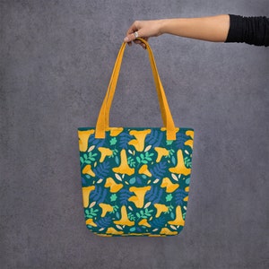 May include: A teal tote bag with a repeating pattern of yellow mushrooms and blue leaves. The bag has yellow straps and is held up by a hand. The bag is made of a durable fabric.