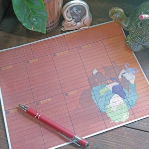 May include: A weekly planner with a reddish-brown background and a cartoon illustration. The planner has sections for each day of the week, with a red pen resting on the surface. A turtle-shaped pencil holder and a cat-shaped picture frame are also visible.