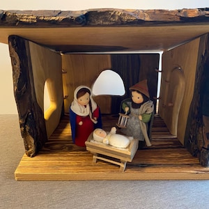 Op de afbeelding: Handgemaakte houten kerststal met figuren van Maria, Jozef en het kindje Jezus. De figuren staan in een rustieke houten constructie met een dak met boomschors. Een traditionele kerstversiering.