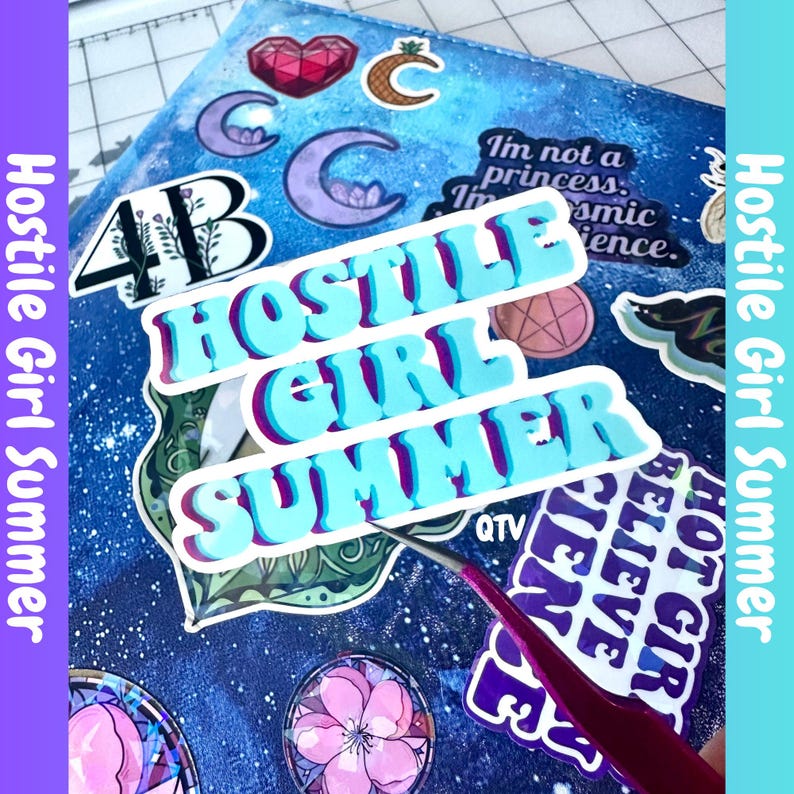 K&ouml;nnte beinhalten: Eine blaue Oberfl&auml;che mit Galaxienmuster, die mit verschiedenen Aufklebern bedeckt ist, darunter ein auff&auml;lliger hellblauer Aufkleber mit dem Text "HOSTILE GIRL SUMMER" und eine d&uuml;nne Metallpinzette, die einen Aufkleber auf die Oberfl&auml;che klebt.