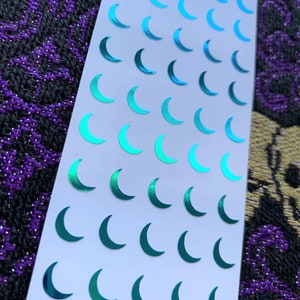 May include: A rectangular white sticker sheet featuring a repeating pattern of small crescent moon stickers. The moons have a metallic, iridescent finish that shifts in color from bright cyan to deep teal across the grid layout.