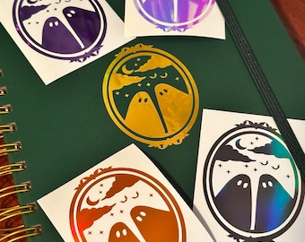 May include: Five oval stickers featuring a pair of ghosts under a crescent moon, clouds, and stars within an ornate frame. The stickers are rendered in various metallic and holographic finishes, including purple, gold, orange, and iridescent blue.