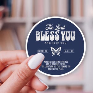May include: A round sticker with a navy blue background and white text. The text reads "The Lord Bless You and Keep You." Includes a butterfly graphic and Bible verse reference. A hand holds the sticker.