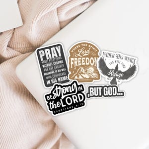May include: A collection of stickers with religious quotes and imagery. The stickers include phrases like "Pray," "Lord Freedom," "Under His Wings," and "Be Strong in the Lord." The stickers are in various shapes and colors, with black, white, and brown tones.
