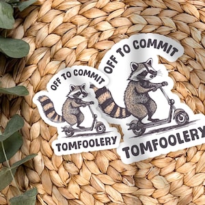 May include: Two stickers featuring a raccoon riding a scooter. The stickers have the text "OFF TO COMMIT" and "TOMFOOLERY". The raccoon is brown and white with a striped tail. The stickers are on a woven, circular surface.