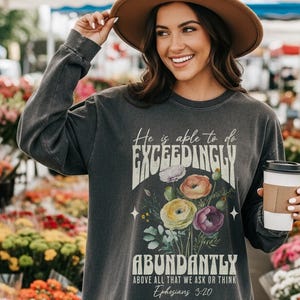 May include: Dark gray long-sleeve shirt with the text "He is able to do EXCEEDINGLY ABUNDANTLY" and a floral design. The text "Ephesians 3:20" is also included. The shirt is made of a soft, comfortable material.