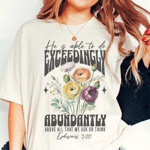 May include: A white t-shirt with a floral design and the text "He is able to do EXCEEDINGLY ABUNDANTLY ABOVE ALL THAT WE ASK OR THINK Ephesians 3:20".