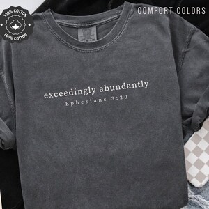 May include: Dark gray long-sleeve shirt with the text "exceedingly abundantly" and "Ephesians 3:20" in white. The shirt is made of 100% cotton and has a Comfort Colors label. The shirt is a crew neck style.
