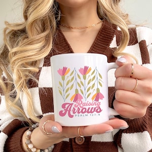 May include: White ceramic mug featuring a floral design with pink and orange flowers and green leaves. The mug has the text "Raising Arrows" and "Psalm 127:4" in pink and white script.