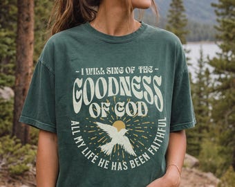 Puede incluir: Una camiseta de manga corta de color verde oscuro con el texto I WILL SING OF THE GOODNESS OF GOD y ALL MY LIFE HE HAS BEEN FAITHFUL en color crema. El gráfico incluye una silueta de pájaro blanca central y un diseño de rayos de sol.