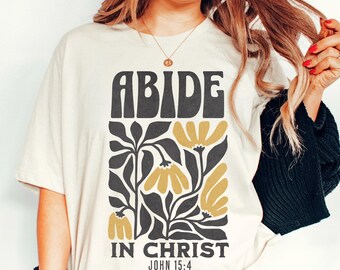 Puede incluir: Una camiseta blanca con un diseño gráfico en negro y amarillo. El gráfico presenta las palabras "ABIDE IN CHRIST" y un diseño floral. El texto "JOHN 15:4" también está incluido.