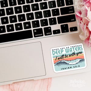 May include: A rectangular sticker on a laptop featuring the text "WHEN YOU GO THROUGH DEEP WATERS I will be with you" with a wave and mountain design. The sticker has a light blue, orange, and teal color scheme. The text "ISAIAH 43:2" is also included.