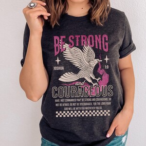 Puede incluir: Una camiseta gris oscura con un gr&aacute;fico rosa de un &aacute;guila con las palabras "Be Strong and Courageous" y un vers&iacute;culo b&iacute;blico de Josu&eacute; 1:9. El texto dice "&iquest;No te he mandado yo? S&eacute; fuerte y valiente; no temas ni te intimides, porque el Se&ntilde;or tu Dios estar&aacute; contigo en dondequiera que vayas."