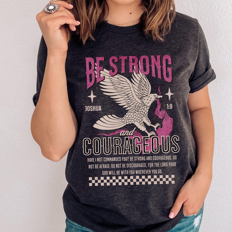 Puede incluir: Una camiseta gris oscura con un gr&aacute;fico rosa de un &aacute;guila con las palabras "Be Strong and Courageous" y un vers&iacute;culo b&iacute;blico de Josu&eacute; 1:9. El texto dice "&iquest;No te he mandado yo? S&eacute; fuerte y valiente; no temas ni te intimides, porque el Se&ntilde;or tu Dios estar&aacute; contigo en dondequiera que vayas."