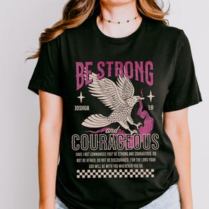 Puede incluir: Camiseta negra con un gr&aacute;fico rosa y blanco de un &aacute;guila con el texto "Be Strong and Courageous" y un vers&iacute;culo b&iacute;blico de Josu&eacute; 1:9.