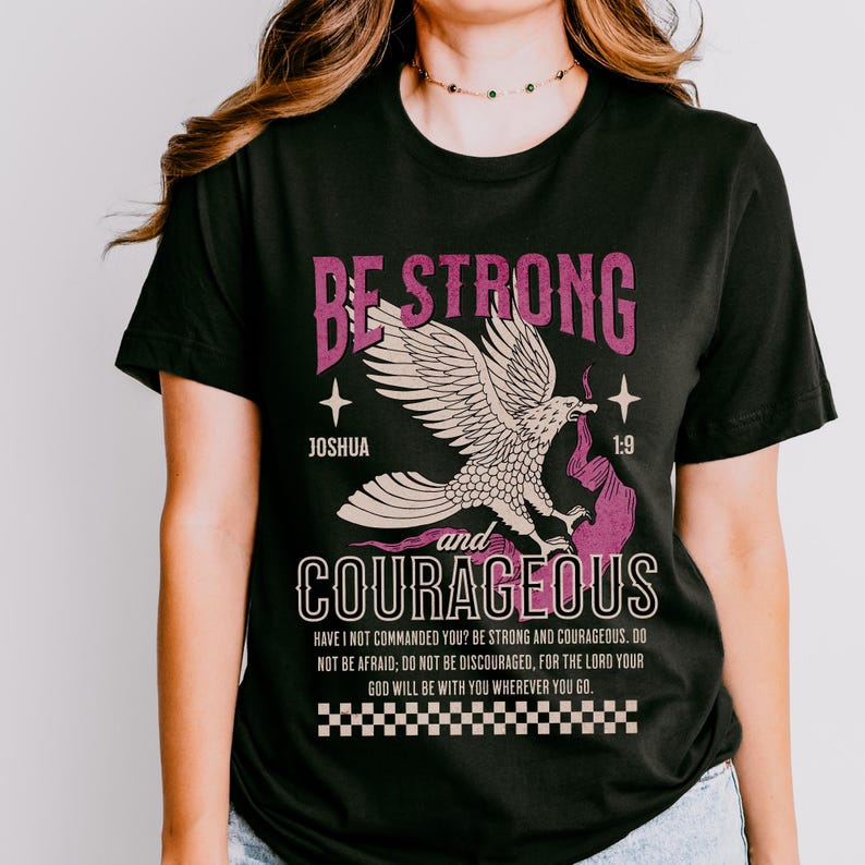 Puede incluir: Camiseta negra con un gr&aacute;fico rosa y blanco de un &aacute;guila con el texto "Be Strong and Courageous" y un vers&iacute;culo b&iacute;blico de Josu&eacute; 1:9.