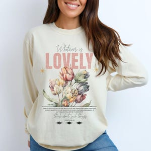May include: Cream-colored long-sleeve sweatshirt with the text "Whatever is LOVELY" in pink. A watercolor floral bouquet of tulips in shades of pink, yellow, and purple is below the text. The sweatshirt also has a quote from Philippians 4:8.