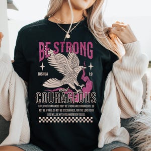 Puede incluir: Camiseta negra con un gr&aacute;fico de &aacute;guila rosa y el texto "Be Strong and Courageous" con el vers&iacute;culo b&iacute;blico Josu&eacute; 1:9. El texto debajo del gr&aacute;fico dice "&iquest;No te he mandado yo? S&eacute; fuerte y valiente; no temas ni te intimides, porque el Se&ntilde;or tu Dios estar&aacute; contigo en dondequiera que vayas."