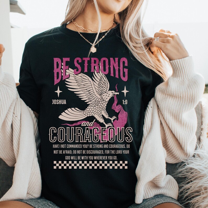 Puede incluir: Camiseta negra con un gr&aacute;fico de &aacute;guila rosa y el texto "Be Strong and Courageous" con el vers&iacute;culo b&iacute;blico Josu&eacute; 1:9. El texto debajo del gr&aacute;fico dice "&iquest;No te he mandado yo? S&eacute; fuerte y valiente; no temas ni te intimides, porque el Se&ntilde;or tu Dios estar&aacute; contigo en dondequiera que vayas."