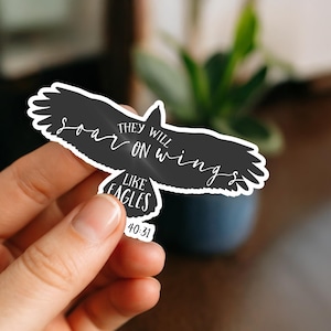 May include: A black sticker in the shape of a soaring eagle with white text that reads "THEY WILL soar ON wings LIKE EAGLES 40:31". The sticker is held by a hand.