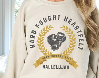 May include: A beige sweatshirt with a graphic design featuring a circle with the text "Hard Fought Heartfelt" and a pair of boxing gloves inside the circle. The text "Been Through Hell" is written on a banner below the circle, and the text "Hallelujah" is written below the banner.