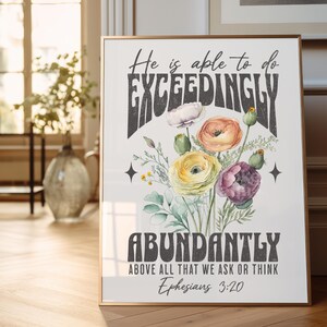 May include: A framed print with a gold border features a watercolor floral arrangement in shades of yellow, orange, and purple. The print includes the text "He is able to do EXCEEDINGLY ABUNDANTLY" and a Bible verse from Ephesians 3:20.
