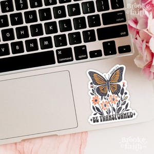 May include: A sticker featuring a butterfly with orange and black wings, surrounded by flowers and leaves. The sticker has the text "Romans 12:2" and "BE TRANSFORMED" on it. The sticker is on a laptop keyboard.