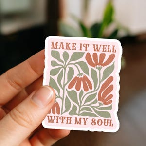 May include: A sticker with a pink border and a floral design. The sticker features the text "MAKE IT WELL WITH MY SOUL" in a brown font. The floral design includes brown flowers and green leaves.