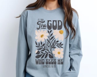 May include: A long-sleeve, light blue shirt with a floral graphic and text. The graphic features white flowers with yellow centers and the words "EL ROI The GOD WHO SEES ME Genesis 16:13" in a black, vintage font.