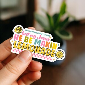 Może przedstawiać: Kolorowa naklejka z napisem "Life gave me some lemons but my Jesus HE BE MAKIN' LEMONADE". Naklejka zawiera ilustrację cytryny oraz r&oacute;żowo-biały wz&oacute;r w kratkę.
