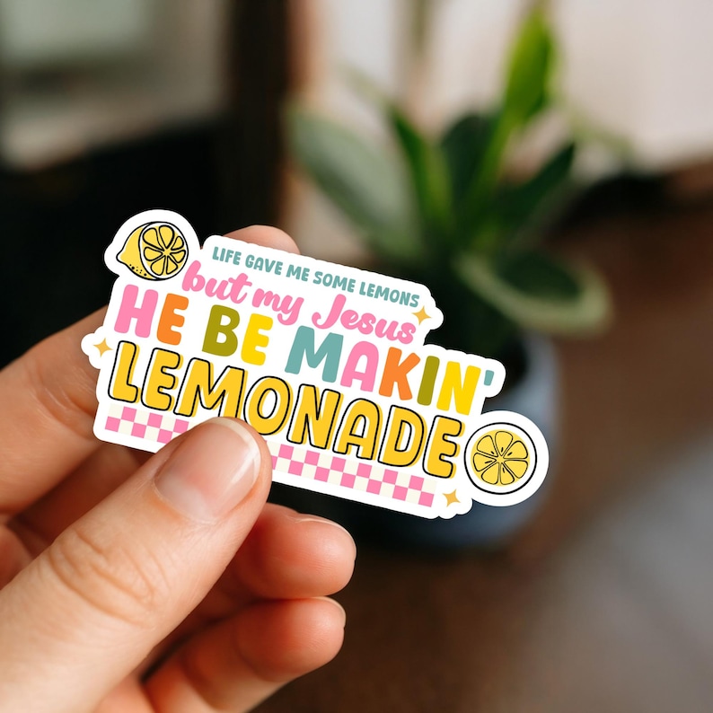 Może przedstawiać: Kolorowa naklejka z napisem "Life gave me some lemons but my Jesus HE BE MAKIN' LEMONADE". Naklejka zawiera ilustrację cytryny oraz r&oacute;żowo-biały wz&oacute;r w kratkę.