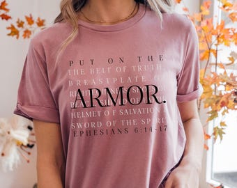 May include: Dusty rose t-shirt with rolled sleeves. The shirt features the word "ARMOR" in large, bold letters, with additional text above and below, including "PUT ON THE BELT OF TRUTH" and "EPHESIANS 6:14-17". The shirt is a solid color.