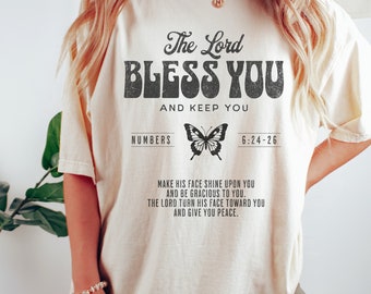 Puede incluir: Una camiseta beige con un gráfico negro que dice "The Lord Bless You and Keep You" con una mariposa y el versículo bíblico Números 6:24-26. El texto debajo del gráfico dice "Make his face shine upon you and be gracious to you. The Lord turn his face toward you and give you peace."