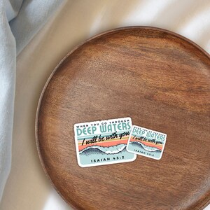 May include: Two stickers on a wooden tray. The stickers have the text "DEEP WATERS I will be with you" and "ISAIAH 43:2". The design includes a mountain and wave graphic with teal, orange, and blue colors.