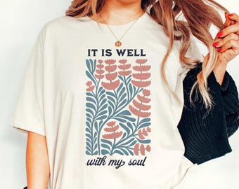 May include: Cream-colored t-shirt featuring the text "IT IS WELL with my soul" above a floral design. The design includes blue leaves and pink flowers. The shirt has short sleeves and a relaxed fit.