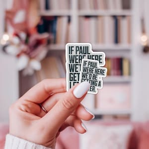May include: A white sticker with black text that reads "IF PAUL WERE HERE WE'D GET IF PAUL WERE HERE WE'D BE PETTING A ER.". The sticker is held up in front of a blurred background.