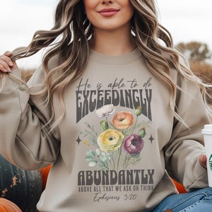 May include: Beige sweatshirt with the text "He is able to do EXCEEDINGLY ABUNDANTLY" and a floral bouquet design in the center. The bouquet features white, yellow, orange, and purple flowers. The Bible verse Ephesians 3:20 is also printed.