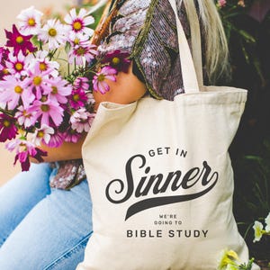 May include: A cream-colored tote bag with the words "GET IN Sinner" in a stylized script, with "WE'RE GOING TO BIBLE STUDY" below. The bag is held by a person carrying a bouquet of pink and purple flowers.