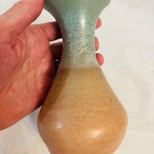 May include: A ceramic vase with a flared rim, featuring a smooth, light tan base that transitions into a textured, mottled sage green glaze on the neck and opening. The vessel has a rounded body and a narrow neck, showcasing a two-tone color palette.