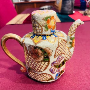May include: A decorative, ornate teapot with a matching lid. The teapot is white with gold accents and features colorful floral designs in orange, pink, and purple. The handle and spout are gold.