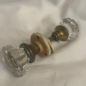 May include: A pair of vintage glass door knobs with brass hardware. The clear glass knobs have a ribbed design. The backplates are painted white and show signs of wear. A classic hardware piece.