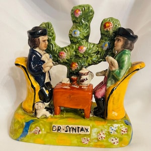 May include: A ceramic figurine depicting two figures seated at a table, with a tree-like structure behind them. The figures are dressed in period clothing, and the table is set with a teapot and cups. The base of the figurine is inscribed with "DR. SYNTAX".