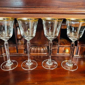 May include: Four clear wine glasses with gold rims. The glasses have a delicate etched design and long stems. They are arranged on a dark wooden surface, suggesting a vintage or antique aesthetic. The glasses are empty and ready for use.