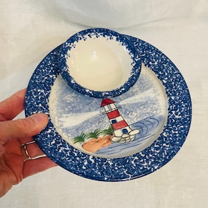 May include: A ceramic plate and bowl set featuring a blue and white speckled rim. The plate displays a hand-painted scene of a red and white lighthouse on a rocky shore with green grass. The bowl is solid cream-colored with a matching speckled blue rim.